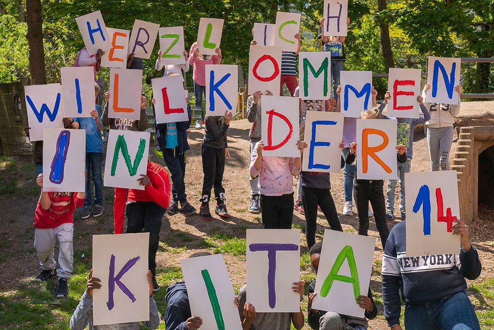 Bild: Kinder halten Schilder hoch, auf denen jeweils ein Buchstabe steht. Daraus ergibt sich folgende Aussage: Herzlich Willkommen in der Kita 14.