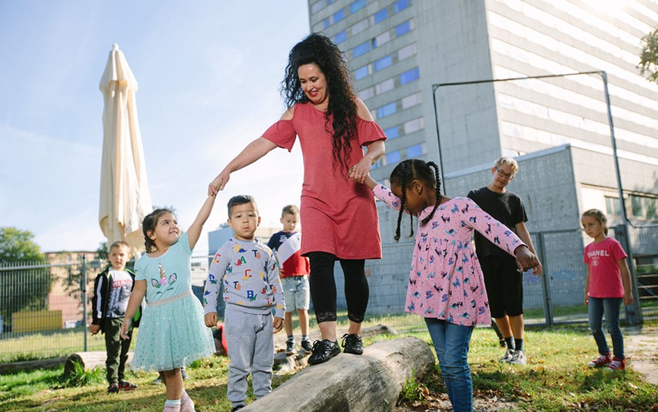 Bild: Eine Erzieherin balanciert auf einem Spielplatz. Sie wird von Kindern geführt. 